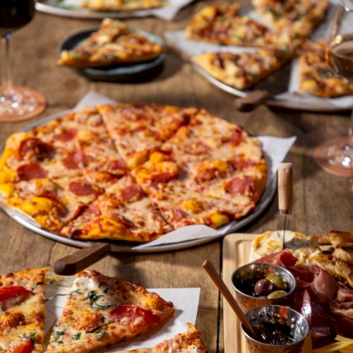 Multiple sliced pizzas with pepperoni on a wooden table, surrounded by bowls of olives and charcuterie, ready to serve.