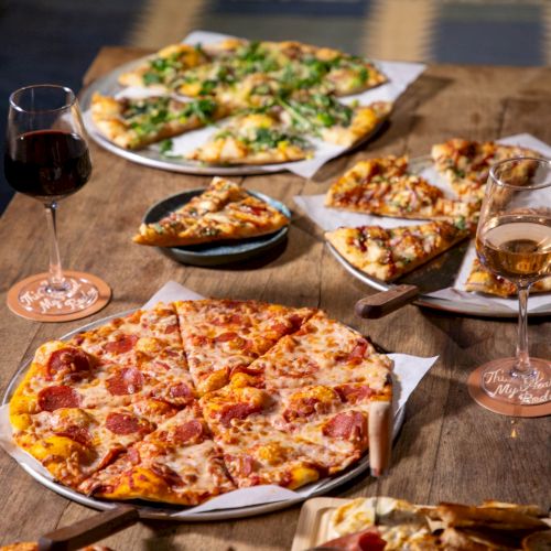 A wooden table piled with cheese and pepperoni pizzas, garlic bread, and two glasses of wine.