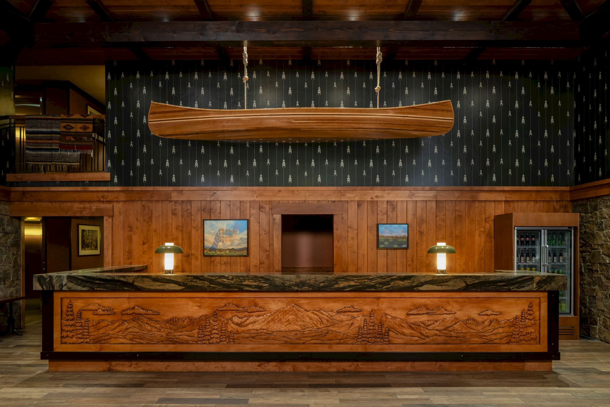 A cozy wooden hotel bar with a carved mural counter, lamps, and a canoe-shaped wooden fixture hanging from the ceiling.