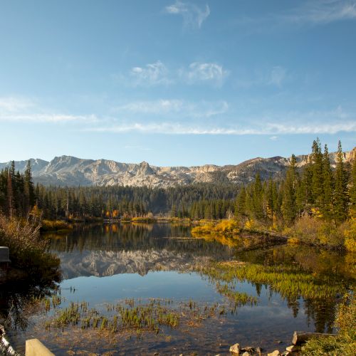 A calm mountain lake reflects autumn trees and rugged peaks under a blue sky with wispy clouds.