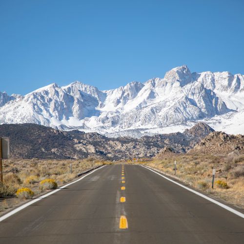 A straight desert road leads toward snow-covered mountains under a clear blue sky, with roadside signs.
