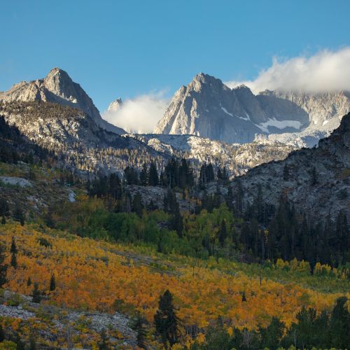 A scenic mountain valley with rocky peaks, autumn-colored forests, and drifting clouds under a clear blue sky.