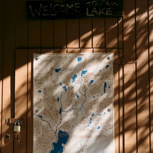 A wooden gate holds a &ldquo;WELCOME LAKE&rdquo; sign and a &ldquo;Sabrina Basin&rdquo; map with shadowed rails and a padlock.