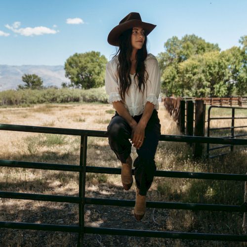 A woman in a cowboy hat sits on a ranch fence in a sunny field with trees and mountains in the distance.