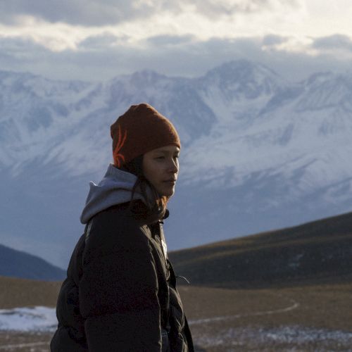 A hiker in winter gear and a knit cap stands against snowy mountains and cloudy skies, looking out quietly.