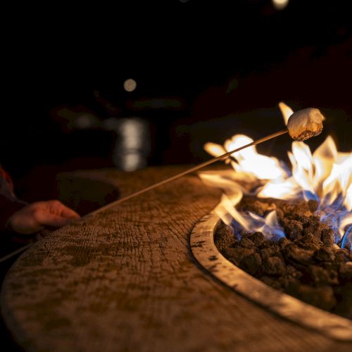 A hand tends a roaring campfire, with a lit log and sparks rising at night.