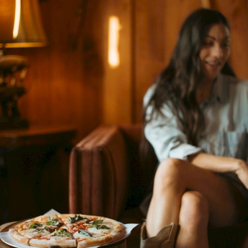 A smiling woman sits in a warm restaurant booth while a plate of pizza rests on the table beside her.