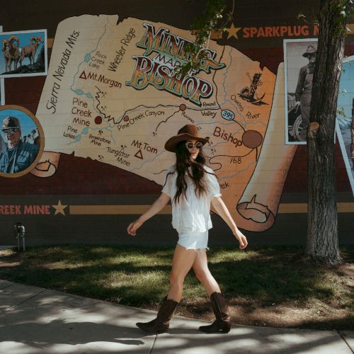 A woman in a cowboy hat and boots poses in front of a colorful Nevada map mural on a building wall.
