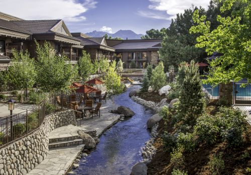 A sunny resort courtyard with a small stream, stone paths, patio seating under red umbrellas, and green buildings with mountains beyond.