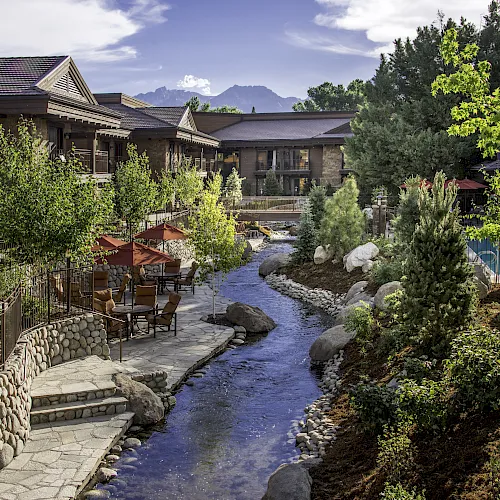 A sunny resort courtyard with a small stream, stone paths, patio seating under red umbrellas, and green buildings with mountains beyond.