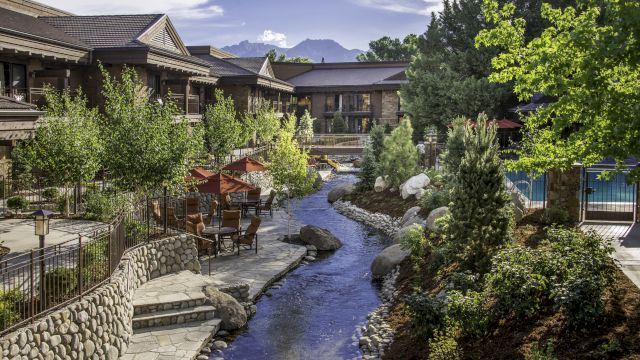 A sunny resort courtyard with a small stream, stone paths, patio seating under red umbrellas, and green buildings with mountains beyond.