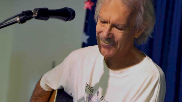 An older man sits and plays an acoustic guitar, singing or speaking into a microphone during a performance.
