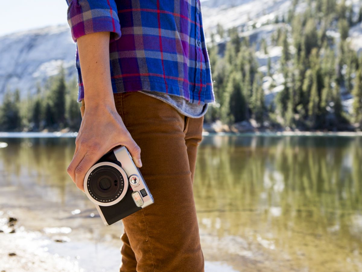 A person in plaid holds a camera by a calm lake, outdoors with trees and mountains in the background.