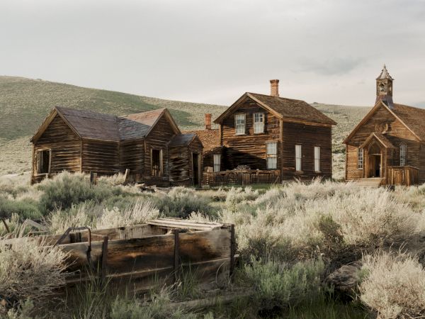An abandoned wooden homestead with weathered buildings and chimneys sits in a grassy desert valley under an overcast sky.