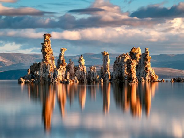 A calm lake with burning, flame-topped rock pillars&mdash;like a fire geyser&mdash;under a cloudy mountain sky.