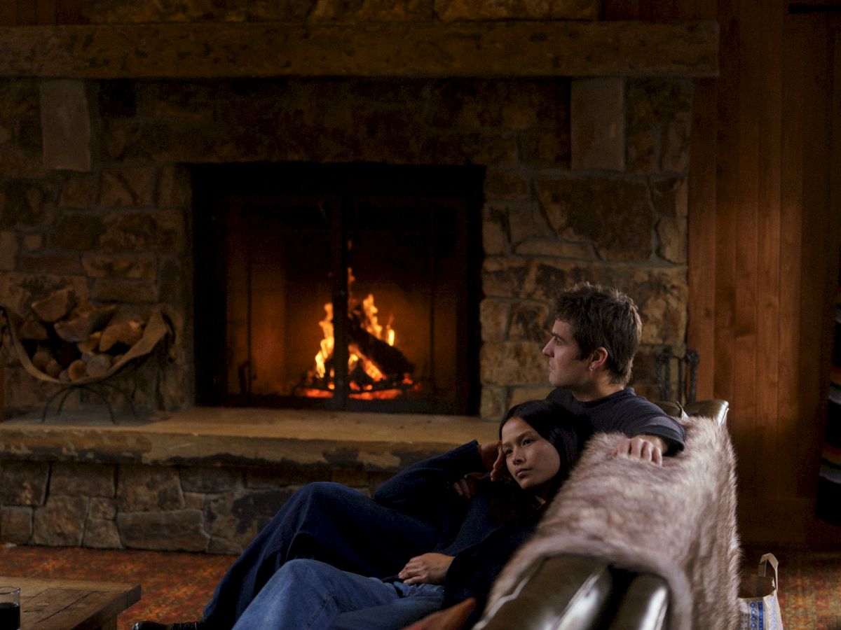 A man cuddles a baby by a cozy stone fireplace while a dog rests nearby on a couch.