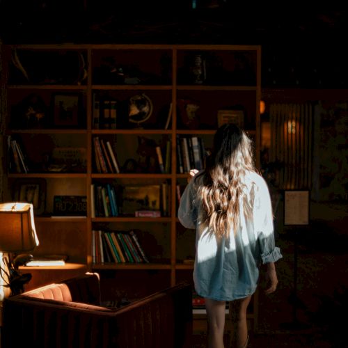 A person with long hair stands in a dim library-like room, with bookshelves, a couch, and warm lamp light illuminating her.