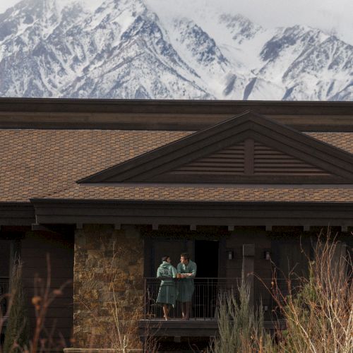 A couple stands on a lodge porch, chatting under snowcapped mountain peaks.