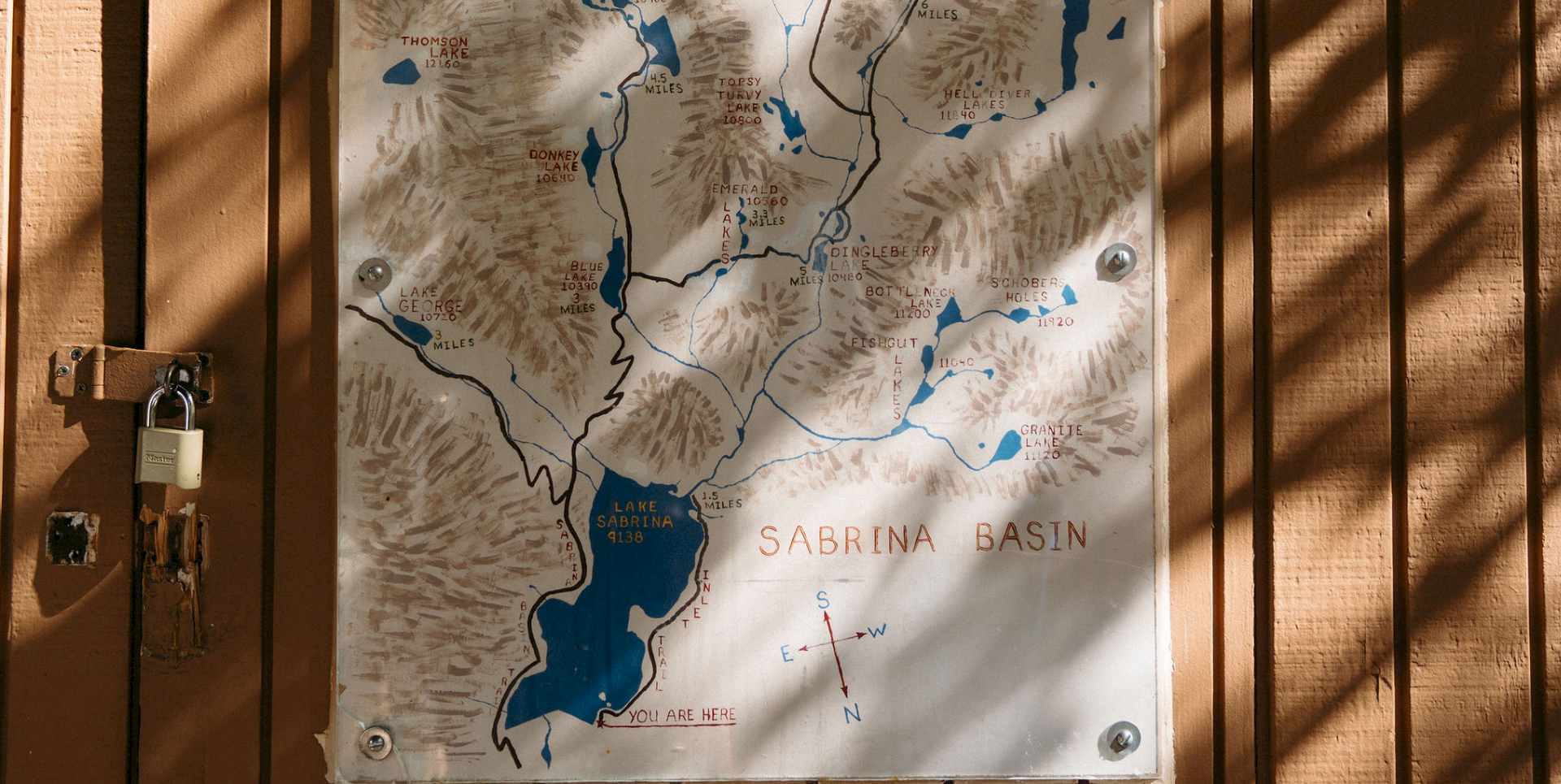 A weathered &ldquo;Sabrina Basin&rdquo; map of lakes and trails, with &ldquo;You are here&rdquo; and metal mounting plates on a wooden wall.