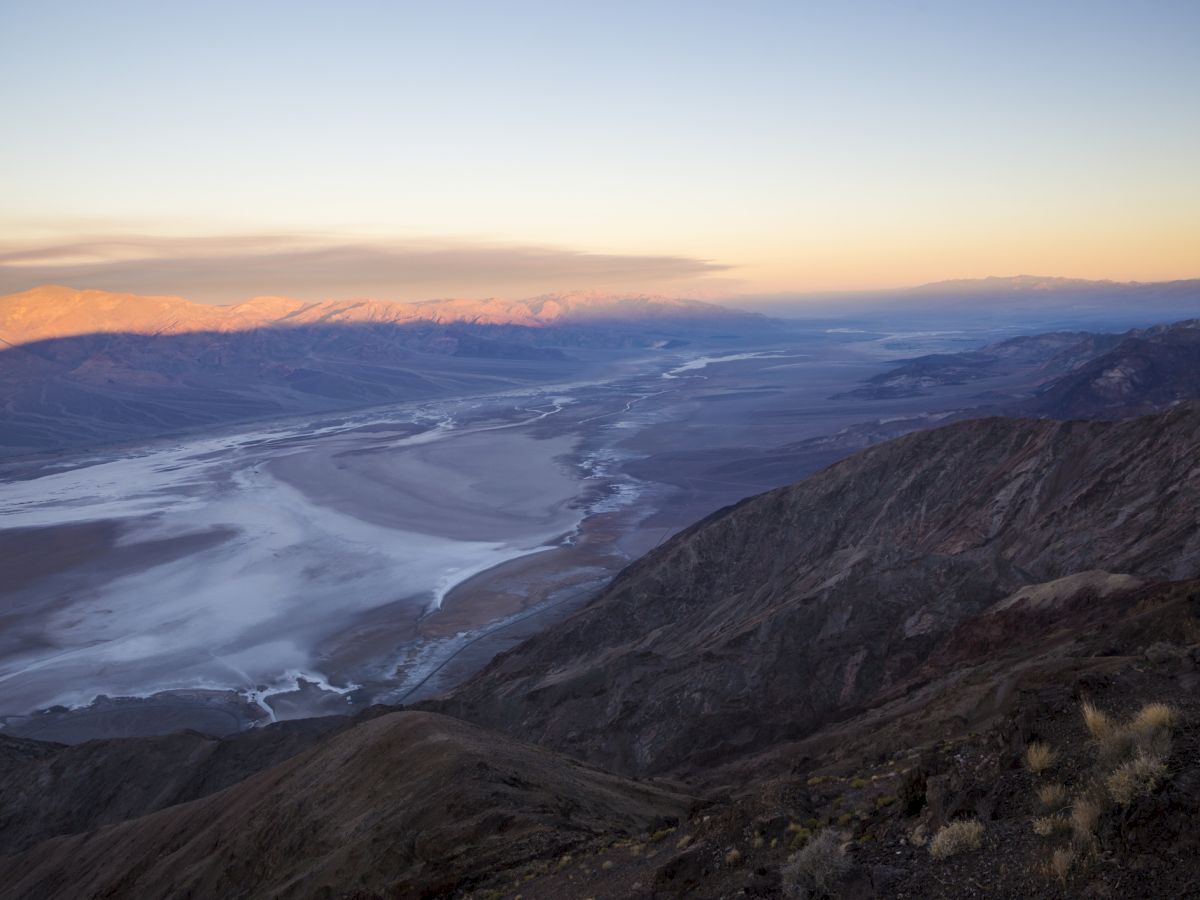 Breathtaking mountain valley at dawn with a winding river/road, vast snowfields, and distant peaks under a pastel sunrise sky.