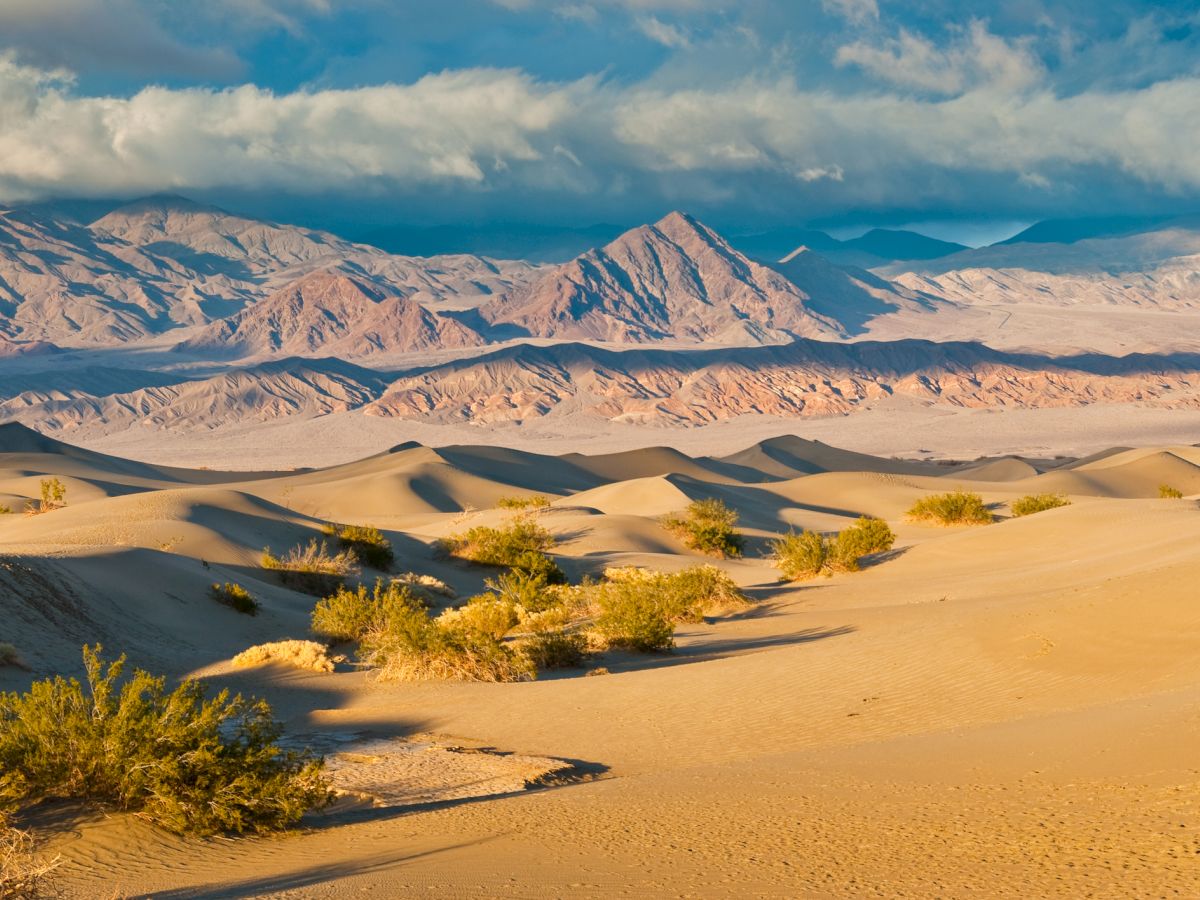 A vast desert of golden sand dunes stretches toward rugged mountain ranges under a bright blue sky with scattered clouds.