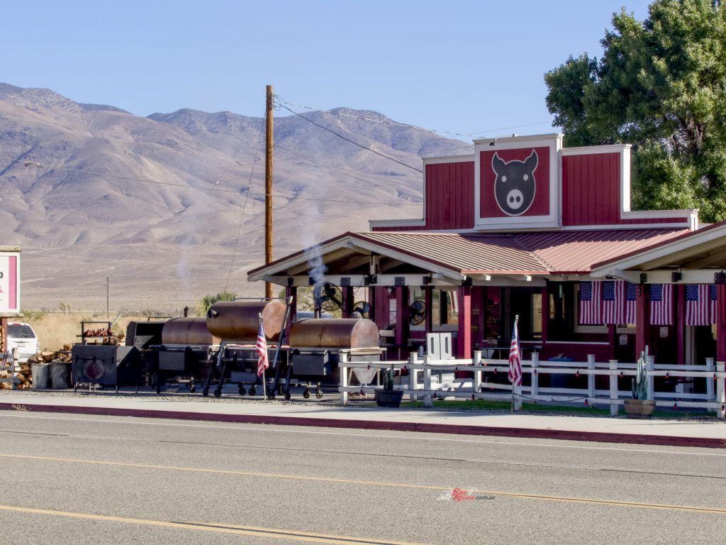 A roadside diner with a red-and-white bunny sign, American flags, and a smoking BBQ pit against desert mountains.