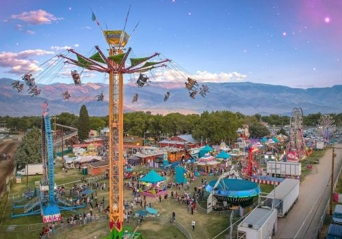 A lively fairground amusement ride lifts daredevils high in the sky above colorful tents and mountains at dusk.