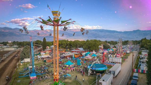 A lively fairground amusement ride lifts daredevils high in the sky above colorful tents and mountains at dusk.