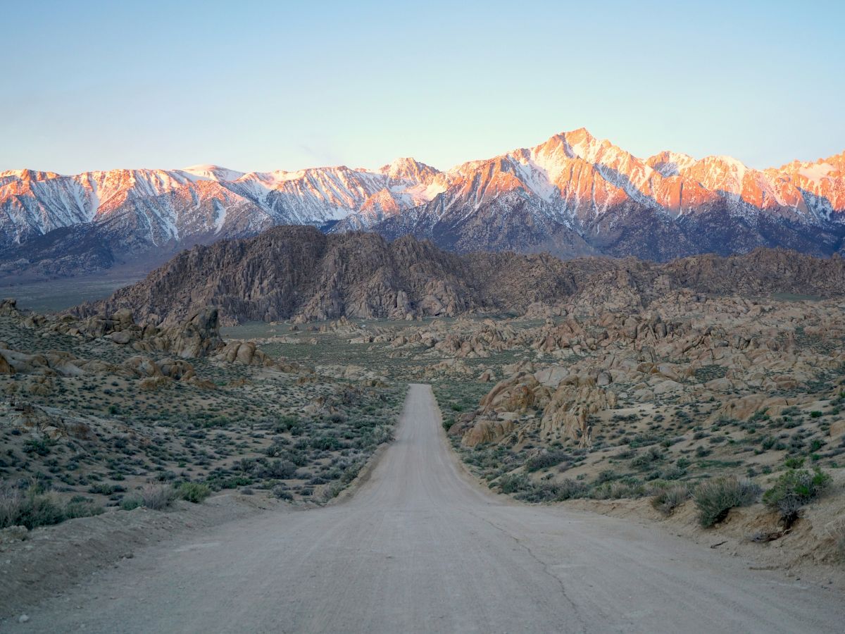 A winding road leads through rocky desert toward snow-dusted mountains glowing at sunrise.