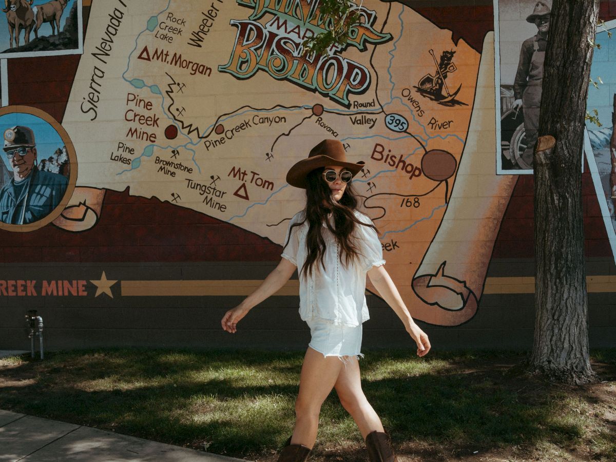 A woman in sunglasses poses in front of a painted mountain-bike map mural on an outdoor wall, with trees nearby.
