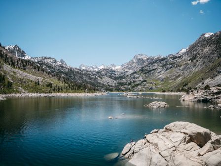 A turquoise alpine lake sits before snowcapped mountain peaks, with rocky shores and scattered boats on a clear day.
