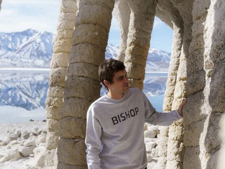 A man poses beside ancient stone arches, framed by snowy mountains and a bright blue sky outside the ruins.