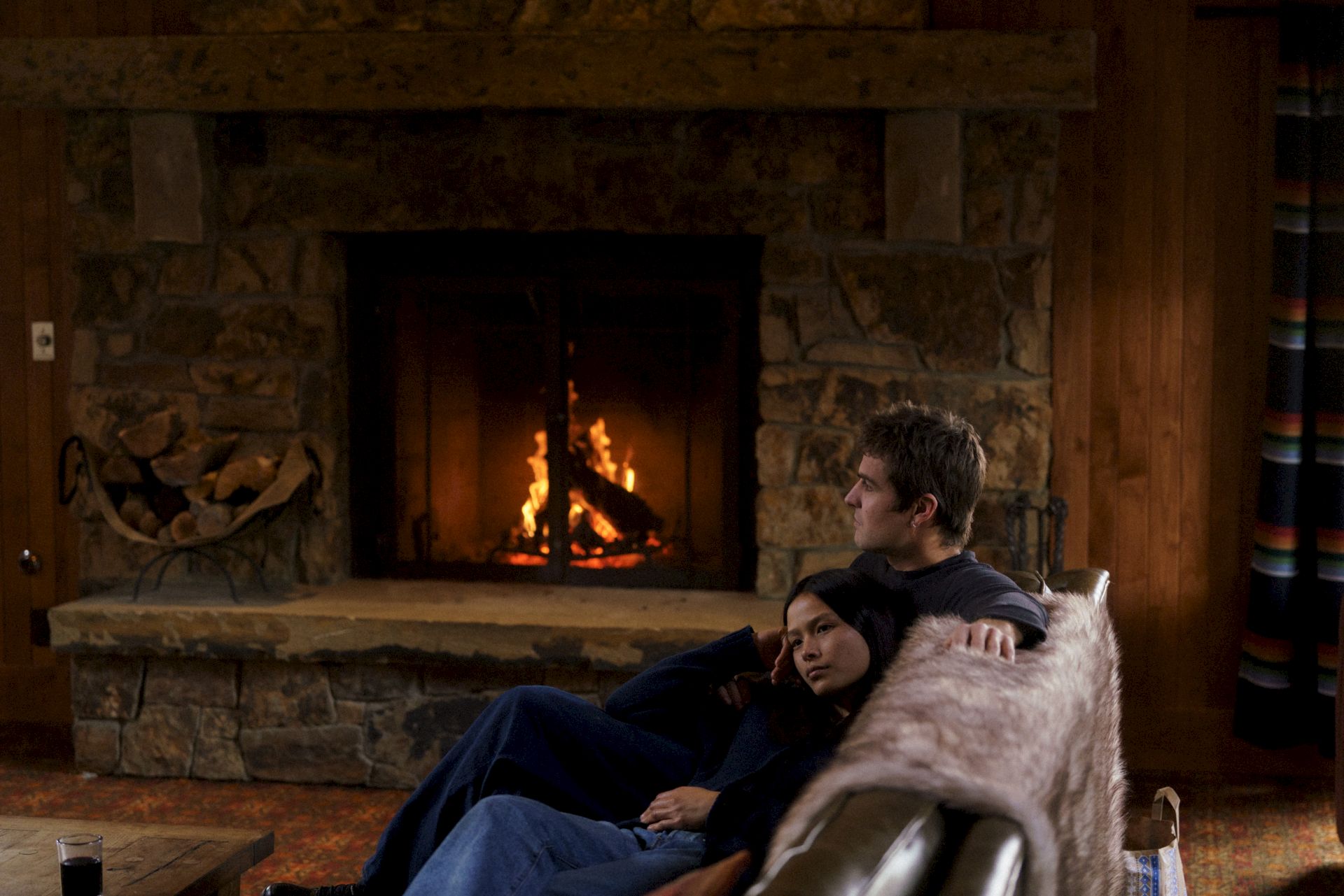 A couple lounges by a stone fireplace with burning logs, while a child rests beside them on a couch.
