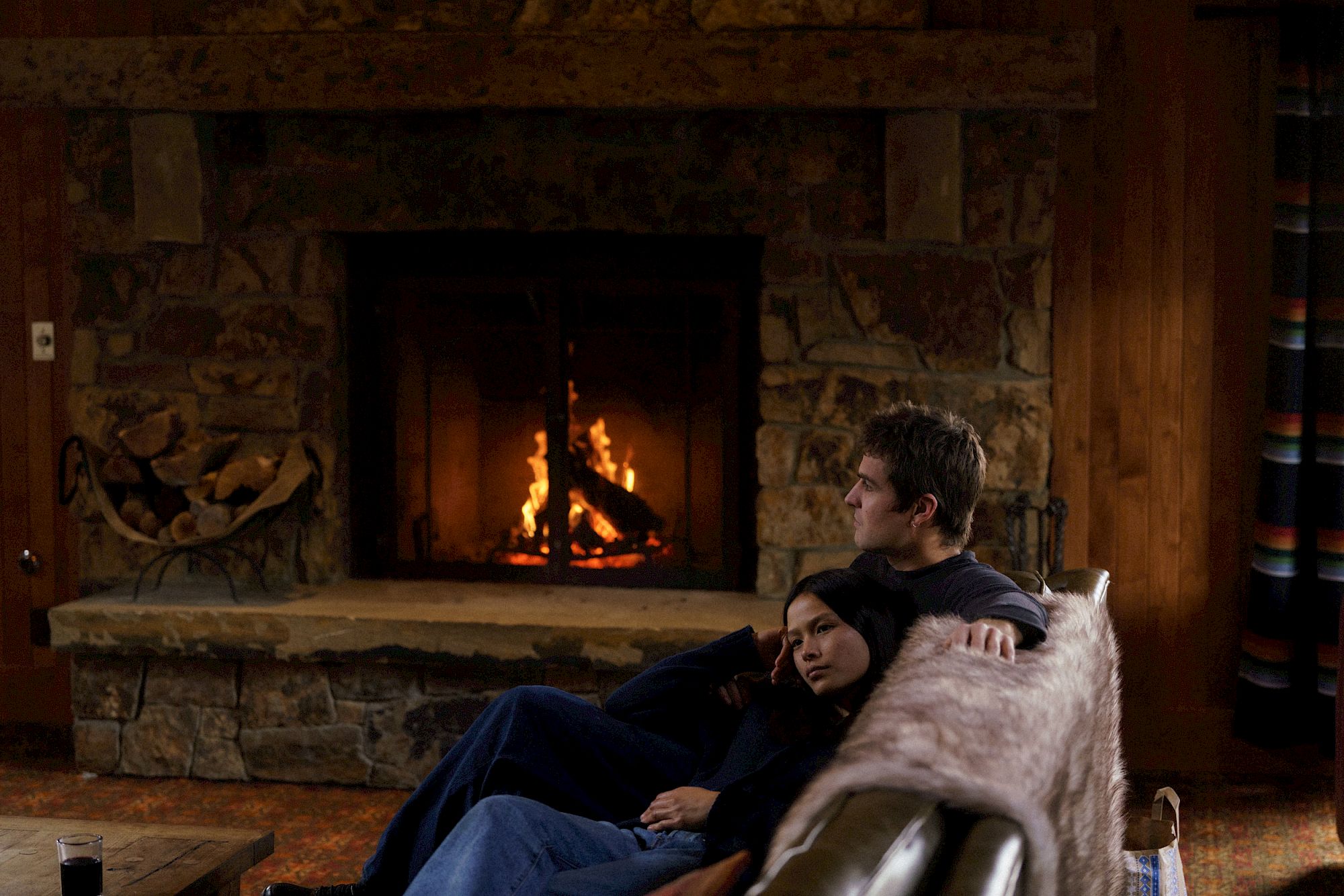 A couple lounges by a stone fireplace with burning logs, while a child rests beside them on a couch.