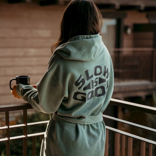 A person in a light green hoodie sits on a balcony, holding a mug and looking out over the street.