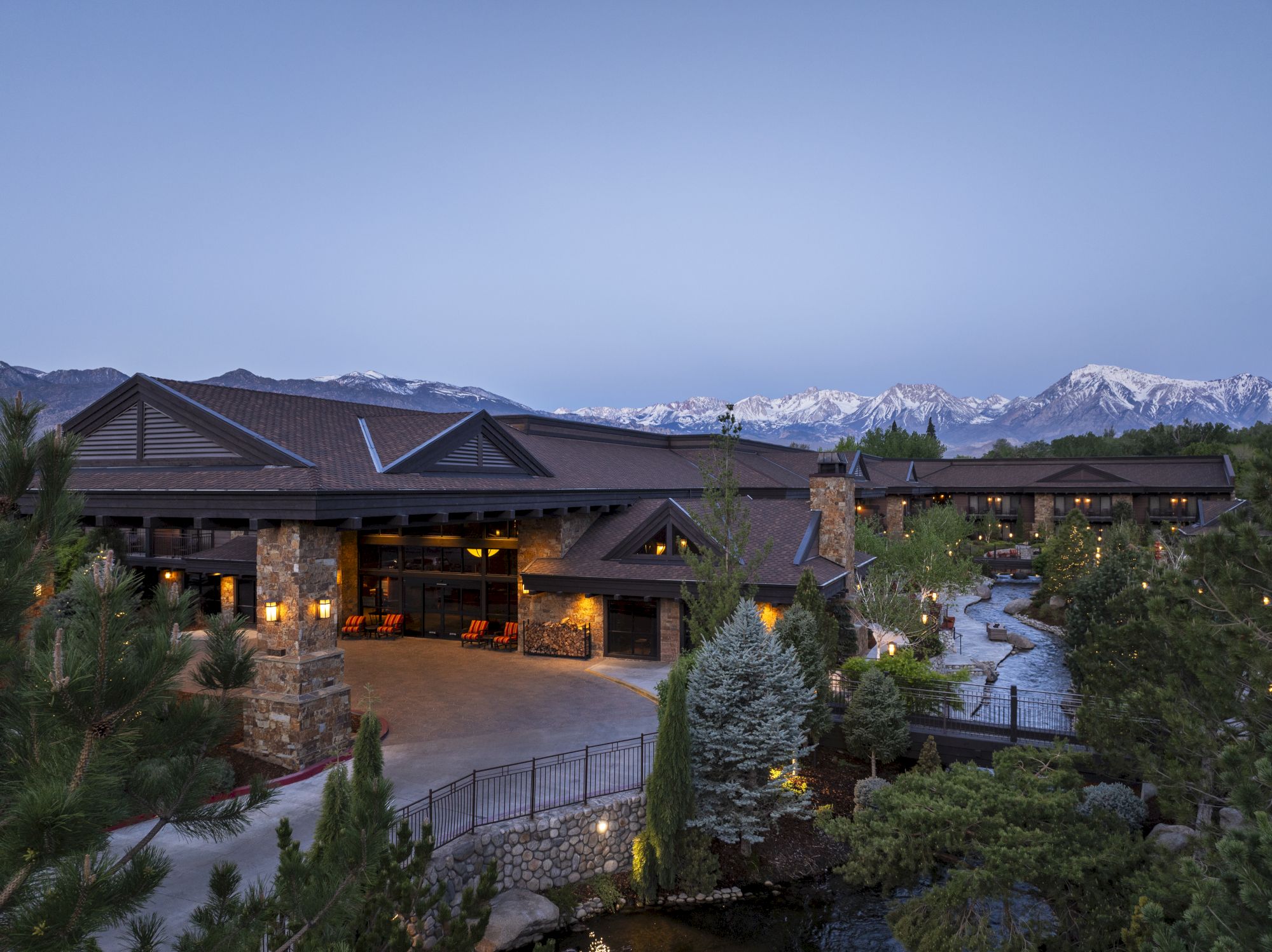 A cozy mountain lodge at dusk with glowing lights and snow-capped peaks in the background.