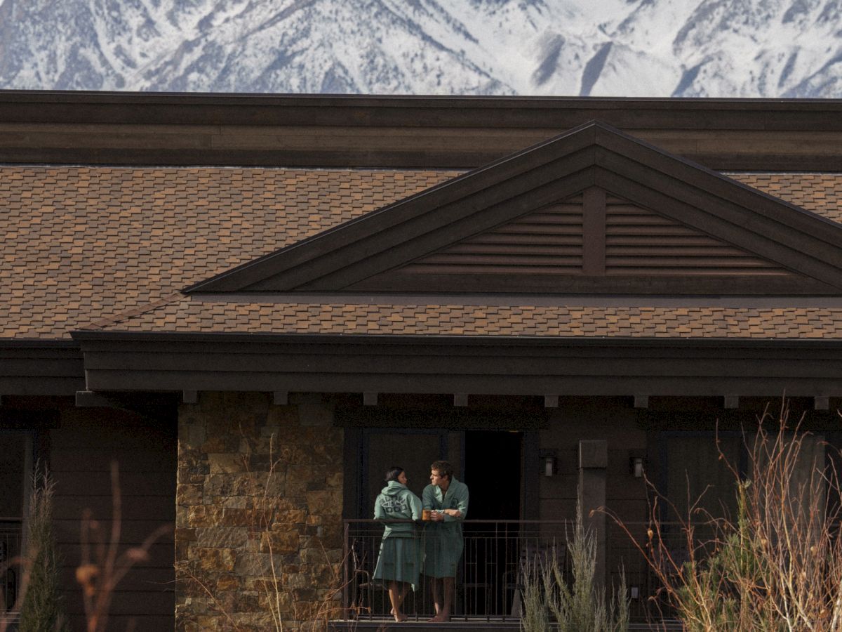 A couple chats on a balcony above snowy peaks, framed by a mountain lodge roof under cloudy skies.
