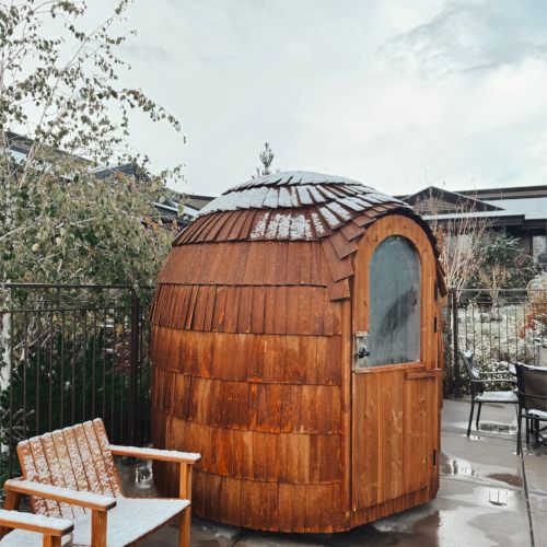 A snowy backyard with a barrel-shaped wooden sauna (or hot tub) and a bench under an overcast sky.