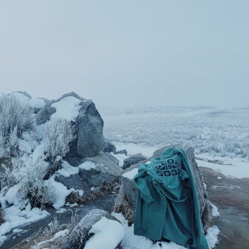 A snowy hillside with frosted rocks and grass, a blue hoodie draped on the stone, and a wide white horizon.