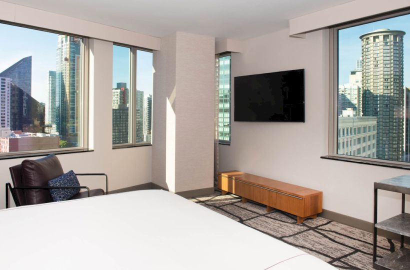 A modern hotel room with large windows showing a city view, featuring a bed, a wall-mounted TV, and a seating area with a chair and table.