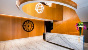A modern lobby features a marble reception desk, large clock on a wooden wall, and contemporary light fixtures with a spiral wooden ceiling accent.