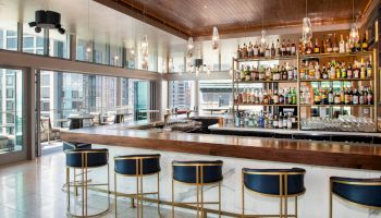 A modern bar with a wooden counter, high stools, and shelves stocked with various bottles. Large windows offer a view of the city.