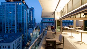 A modern cityscape at dusk featuring tall buildings and a furnished balcony with tables and chairs, overlooking a street.