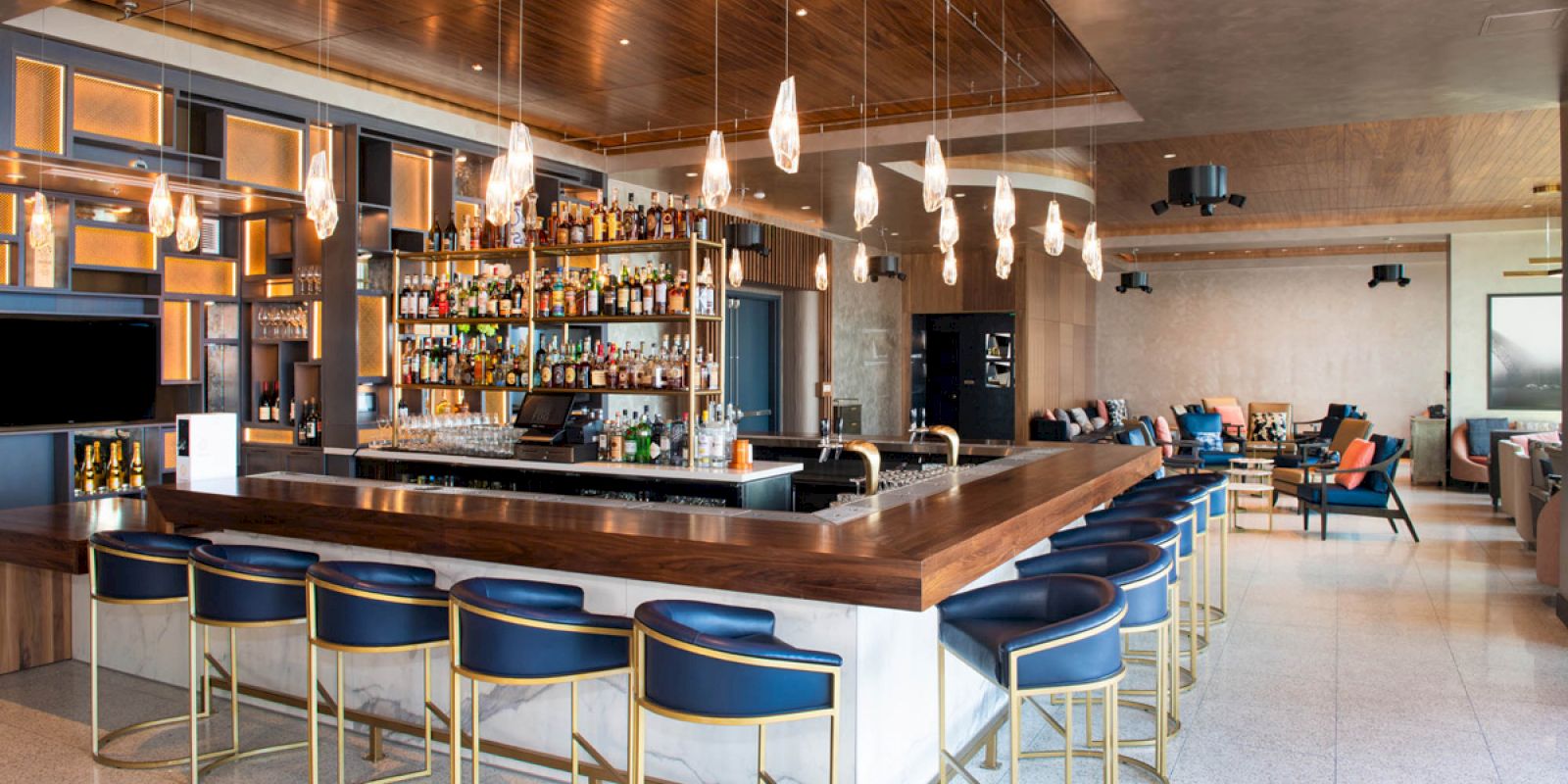 A modern bar with a wooden countertop, blue cushioned barstools, hanging light fixtures, and a well-stocked back bar area.