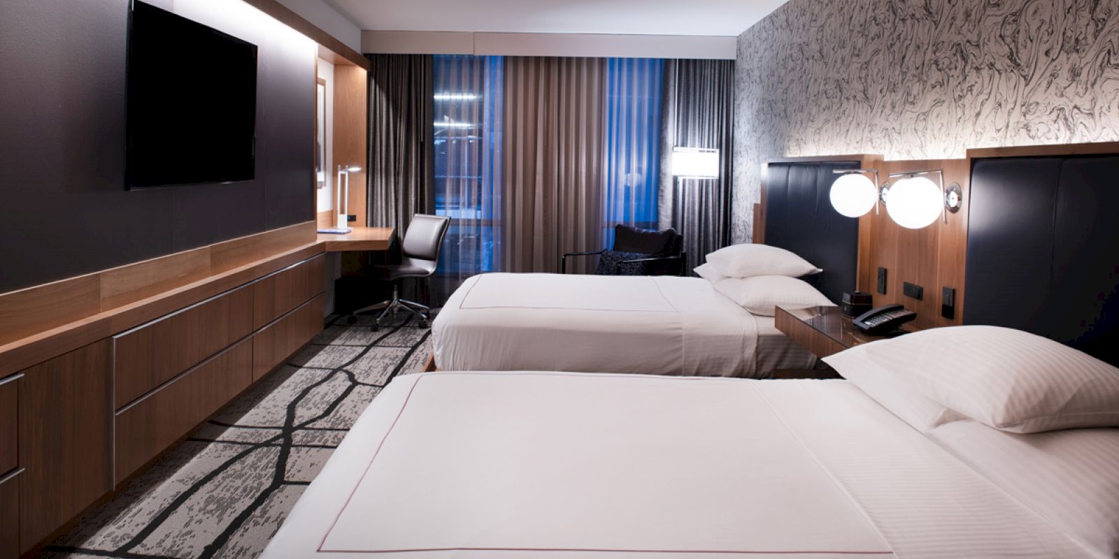 A modern hotel room with two neatly made beds, a wall-mounted TV, a desk and chair by the window, and contemporary design accents.