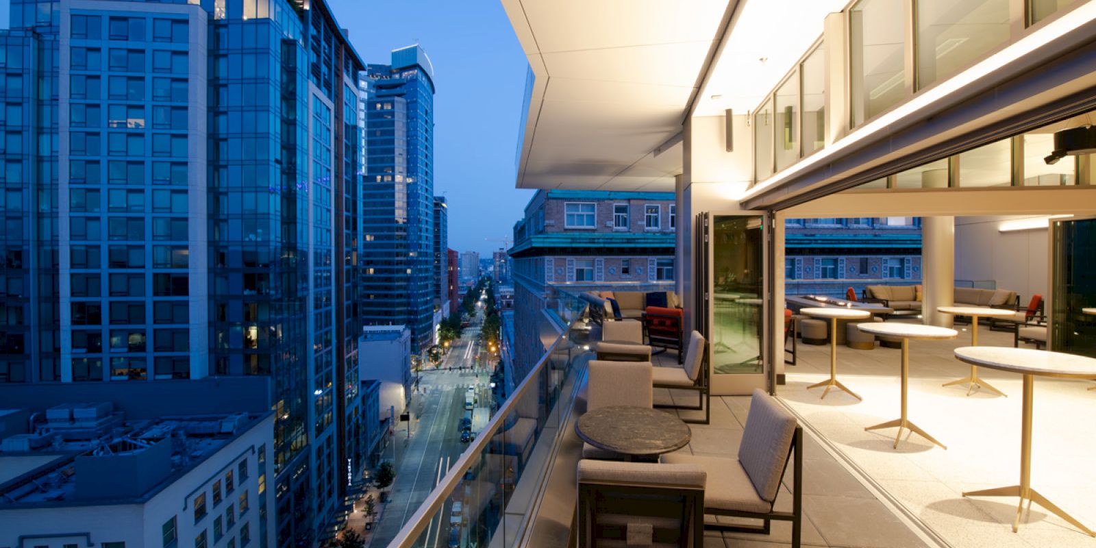 A modern cityscape at dusk with tall buildings and a stylish outdoor terrace featuring tables and chairs overlooking the urban scene.