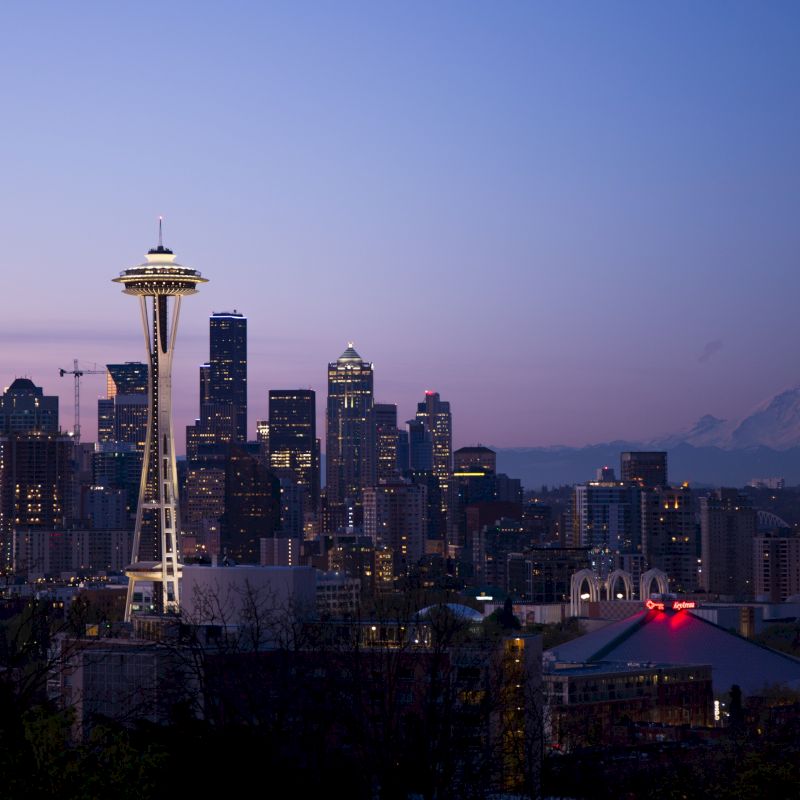 The image shows the Seattle skyline at dusk, featuring the Space Needle with Mount Rainier visible in the background. The sky is transitioning to night.