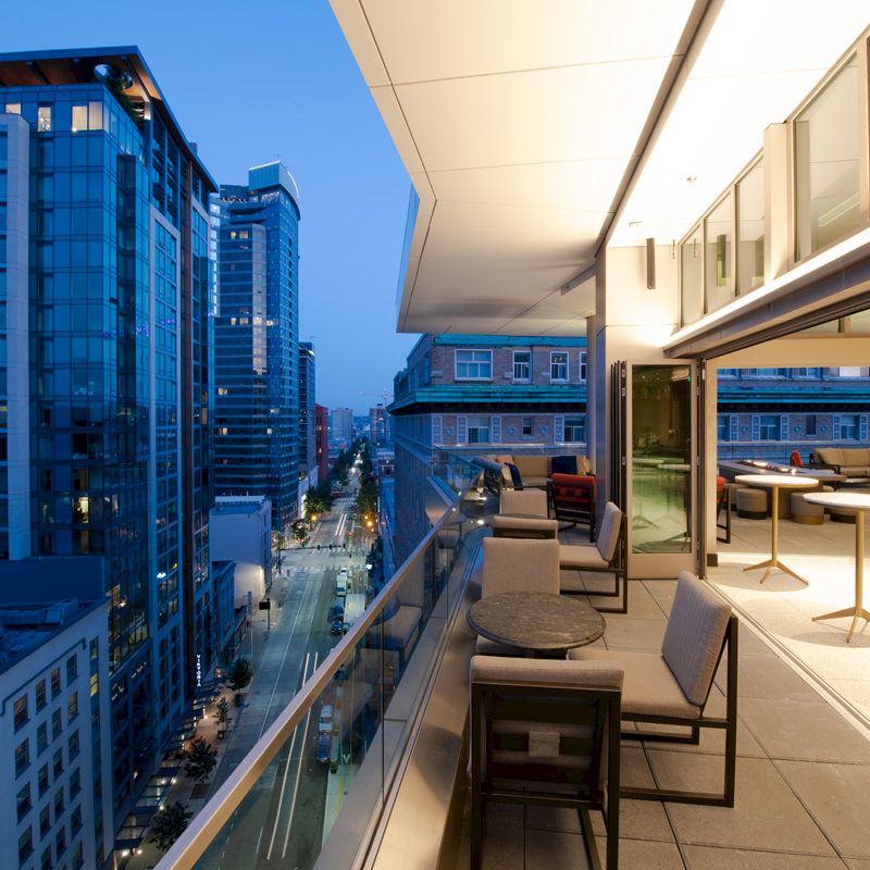 The image shows a modern outdoor patio with tables and chairs, overlooking a cityscape with tall buildings at dusk.