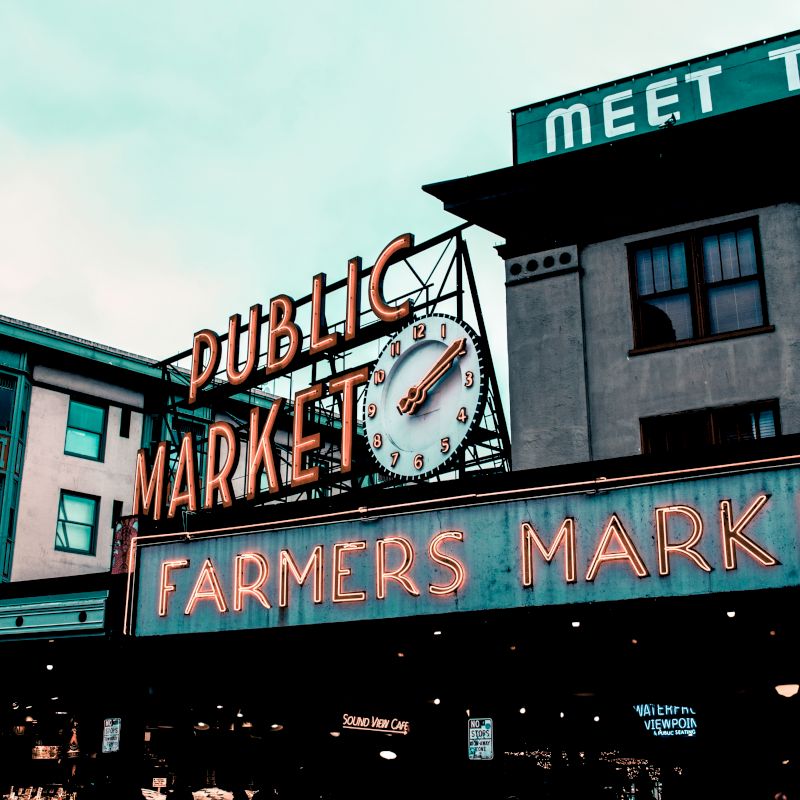 A view of a public market with a neon sign reading "FARMERS MARKET" and a clock above. The surrounding buildings complete the scene.