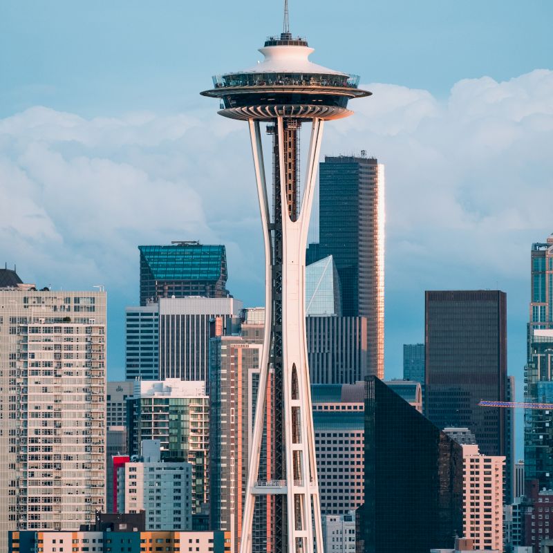 The image shows the Space Needle against a backdrop of modern skyscrapers and buildings under a partly cloudy sky.